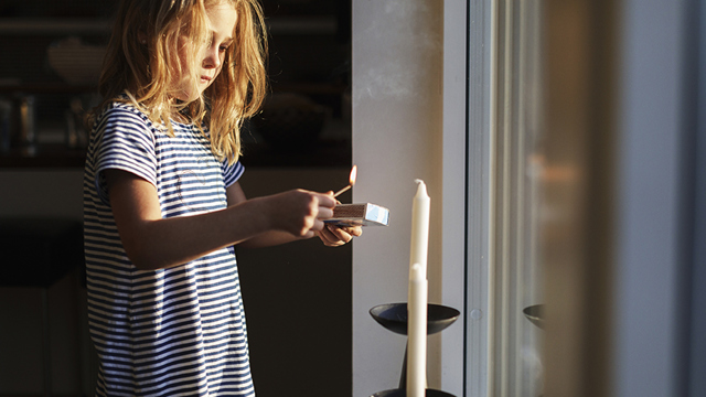 En yngre barn i närbild står bredvid ett fönster och tänder ett stearinljus med en tändsticka. 